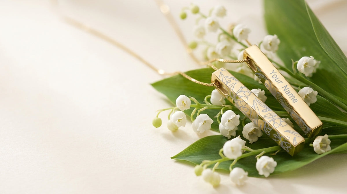 Two gold rectangular pendant necklaces with floral engravings and the text 'Your Name' displayed on a soft cream background, styled with green leaves and white lily of the valley flowers.